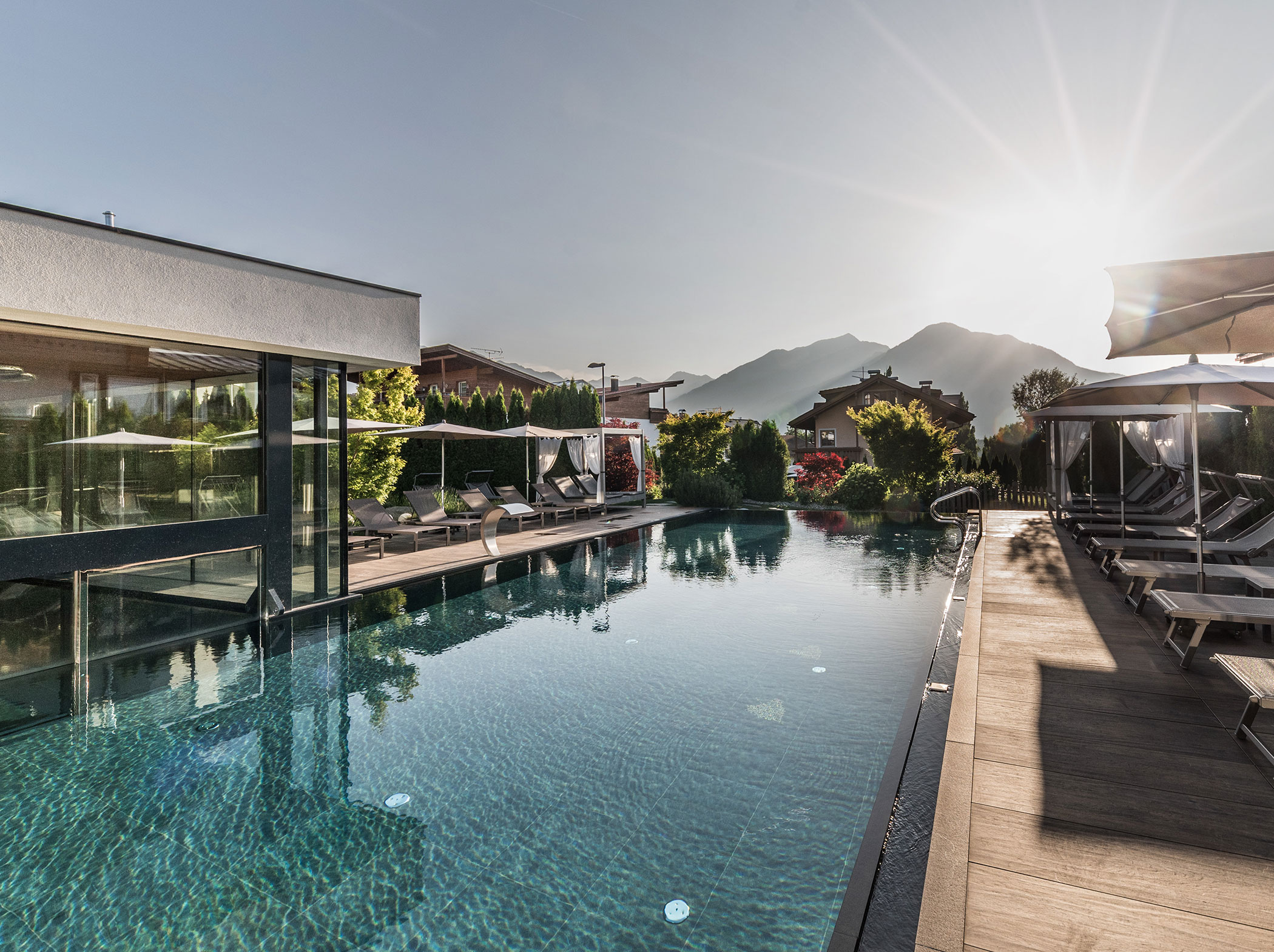 Außenpool mit Liegestühlen neben einem modernen Gebäude, Berge und Sonne im Hintergrund. - Hotel Sun