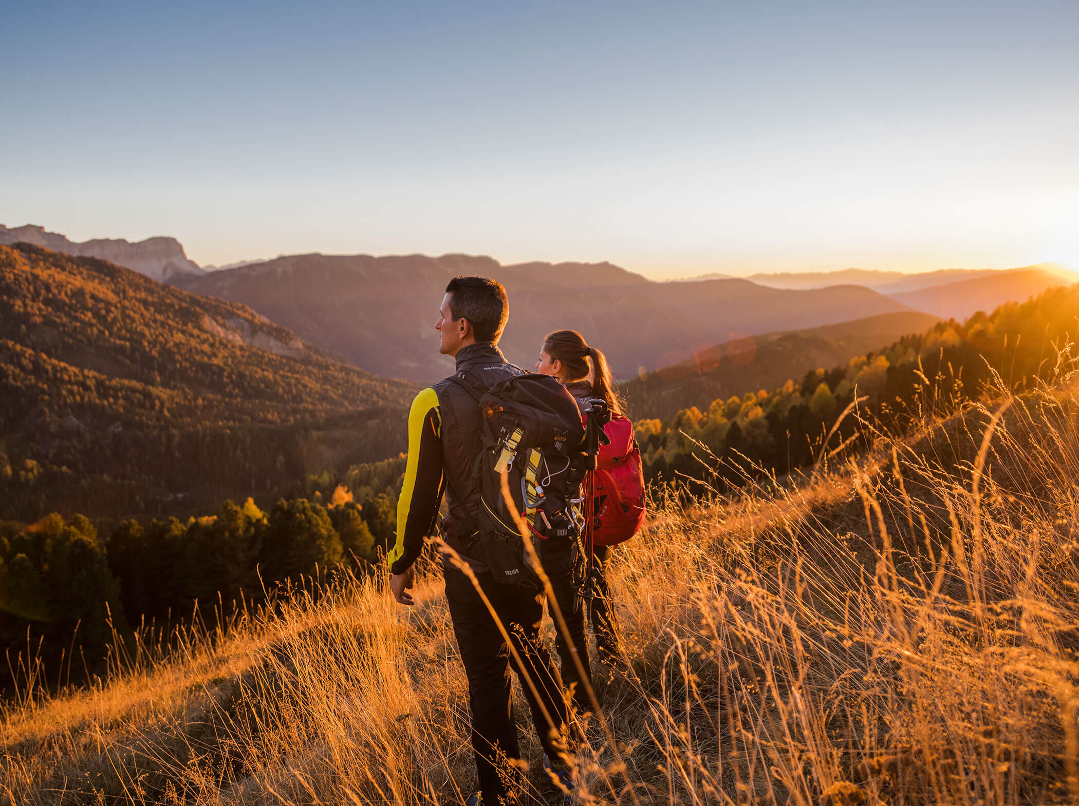 Zwei Wanderer mit Rucksäcken stehen auf einem grasbewachsenen Hügel und betrachten die Berge bei Sonnenuntergang. - Hotel Sun