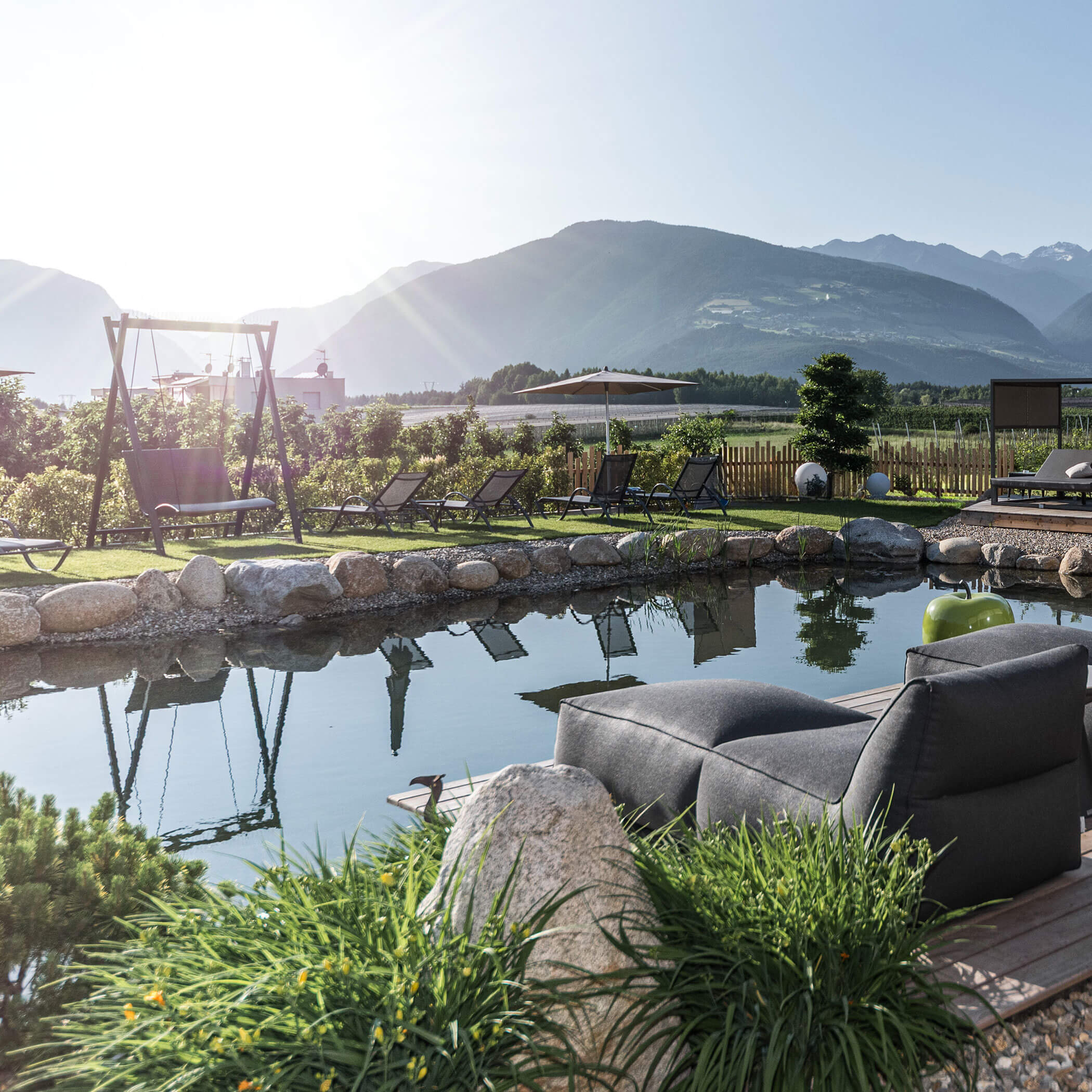 Eine ruhige Lounge im Freien mit Stühlen an einem Teich, Bergen im Hintergrund und hellem Sonnenlicht. - Hotel Sun