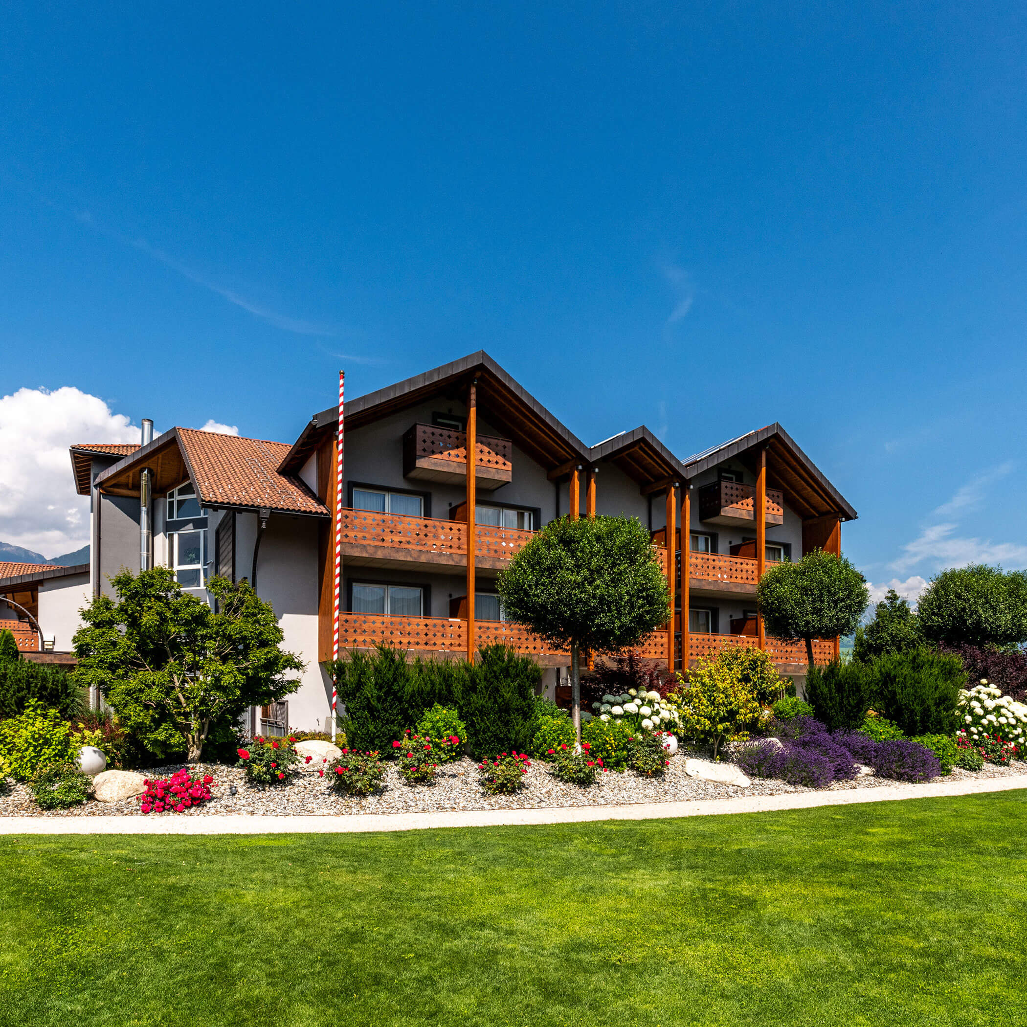 Three-story modern building with wooden balconies, lush landscaping, and a manicured green lawn under a blue sky. - Hotel Sun
