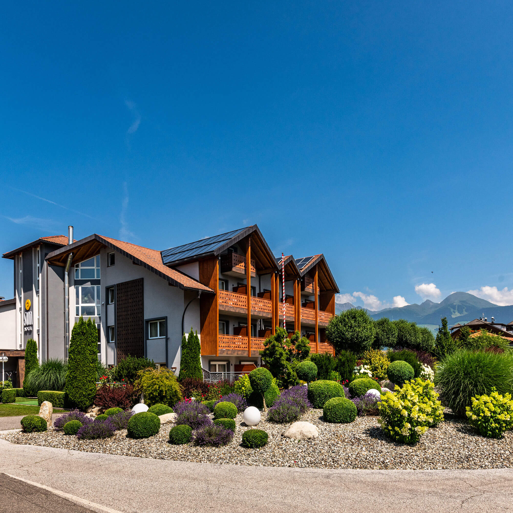 Ein modernes Hotel im Chalet-Stil mit angelegten Gärten, unter blauem Himmel und mit Bergen im Hintergrund. - Hotel Sun