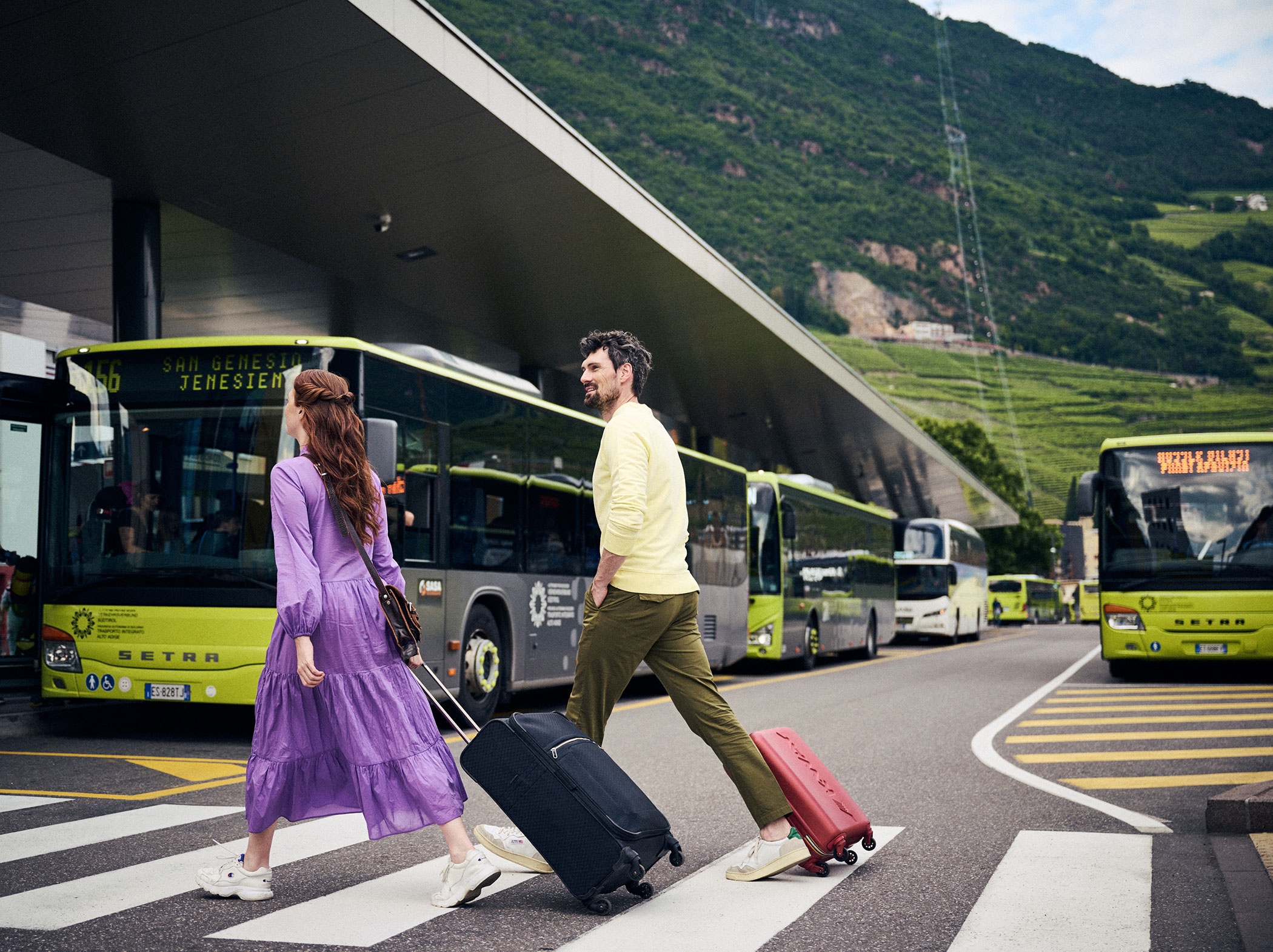 Zwei Personen mit Koffern überqueren eine Straße an einem Busbahnhof mit grünen Bergen im Hintergrund. - Hotel Sun