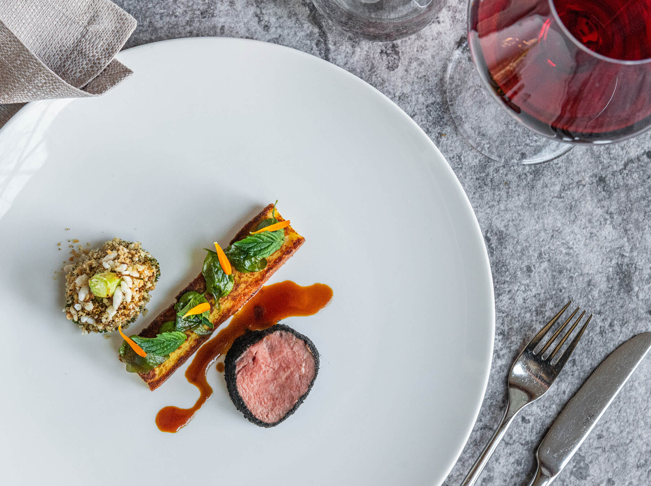 Gourmetgericht mit Fleisch, Gemüse, Beilage, Soße und einem Glas Rotwein auf einem grauen Tisch. - Hotel Sun