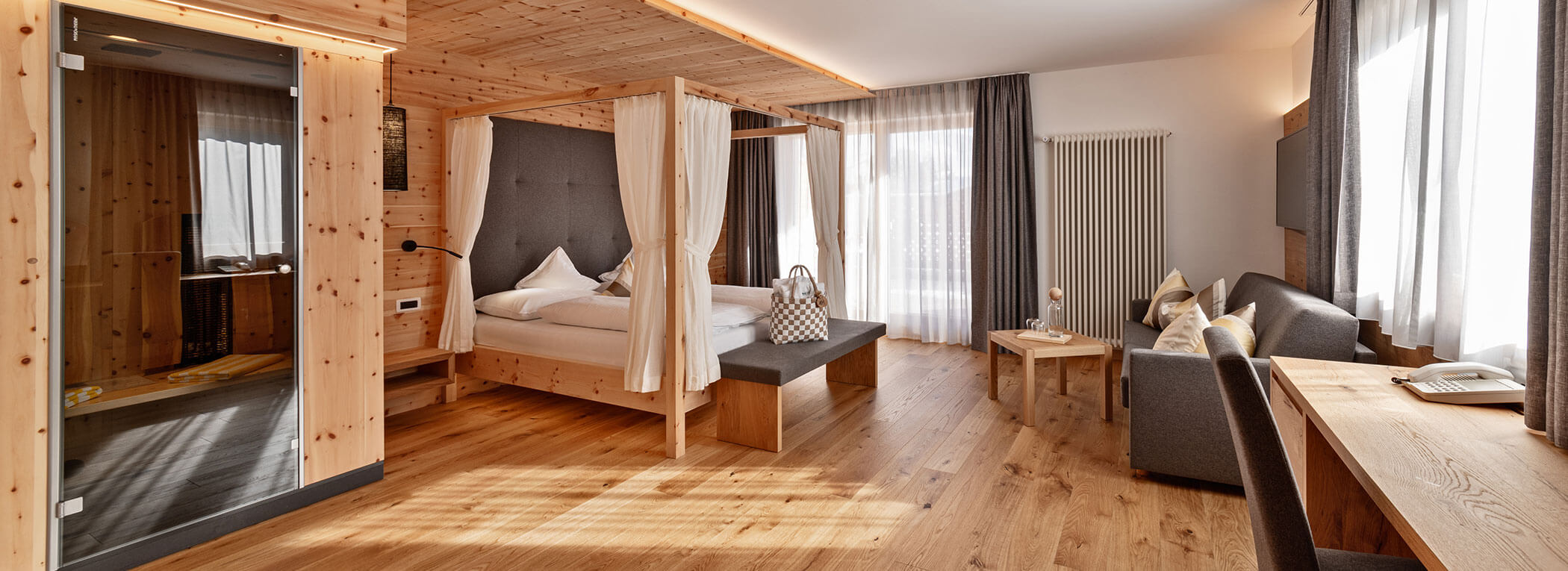 Geräumiges Hotelzimmer aus Holz mit Himmelbett, Sofa, Schreibtisch und großen Fenstern, die viel Tageslicht hereinlassen. - Hotel Sun