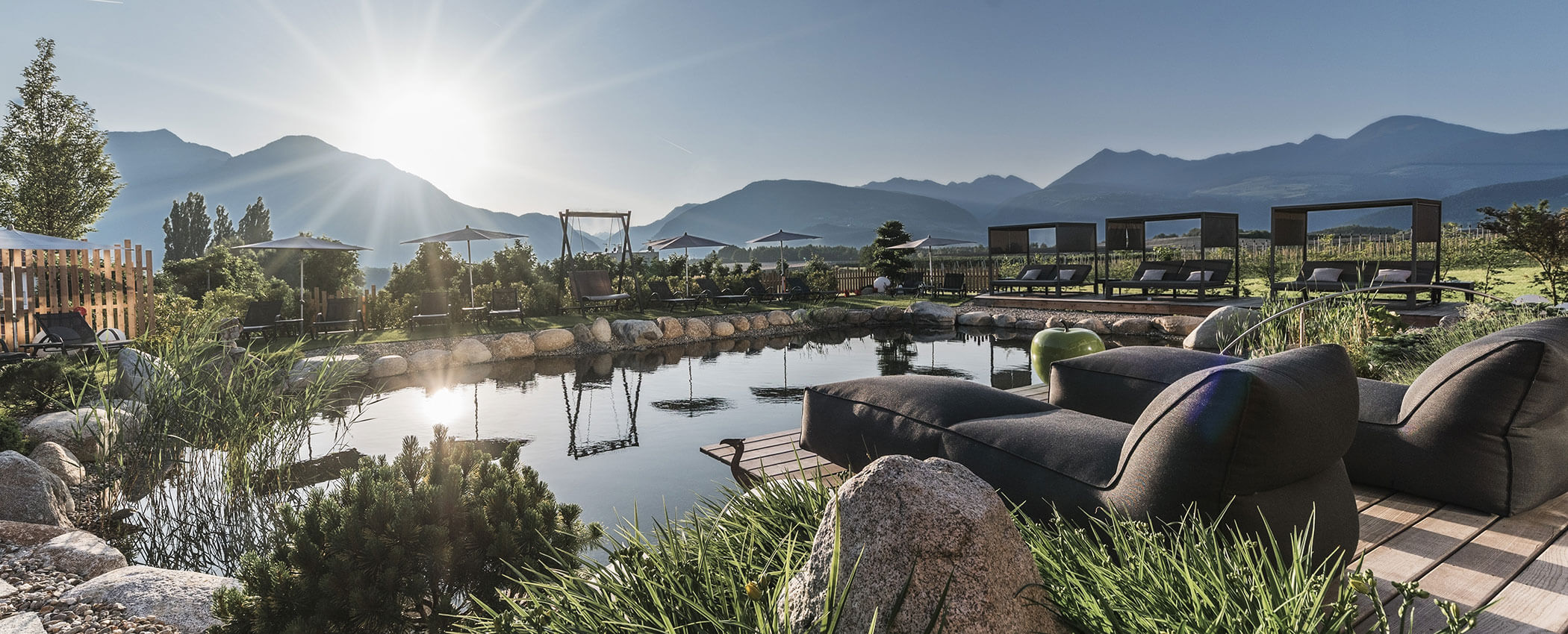 Lounge chairs by a pond with mountain views at sunrise in a landscaped outdoor garden setting. - Hotel Sun