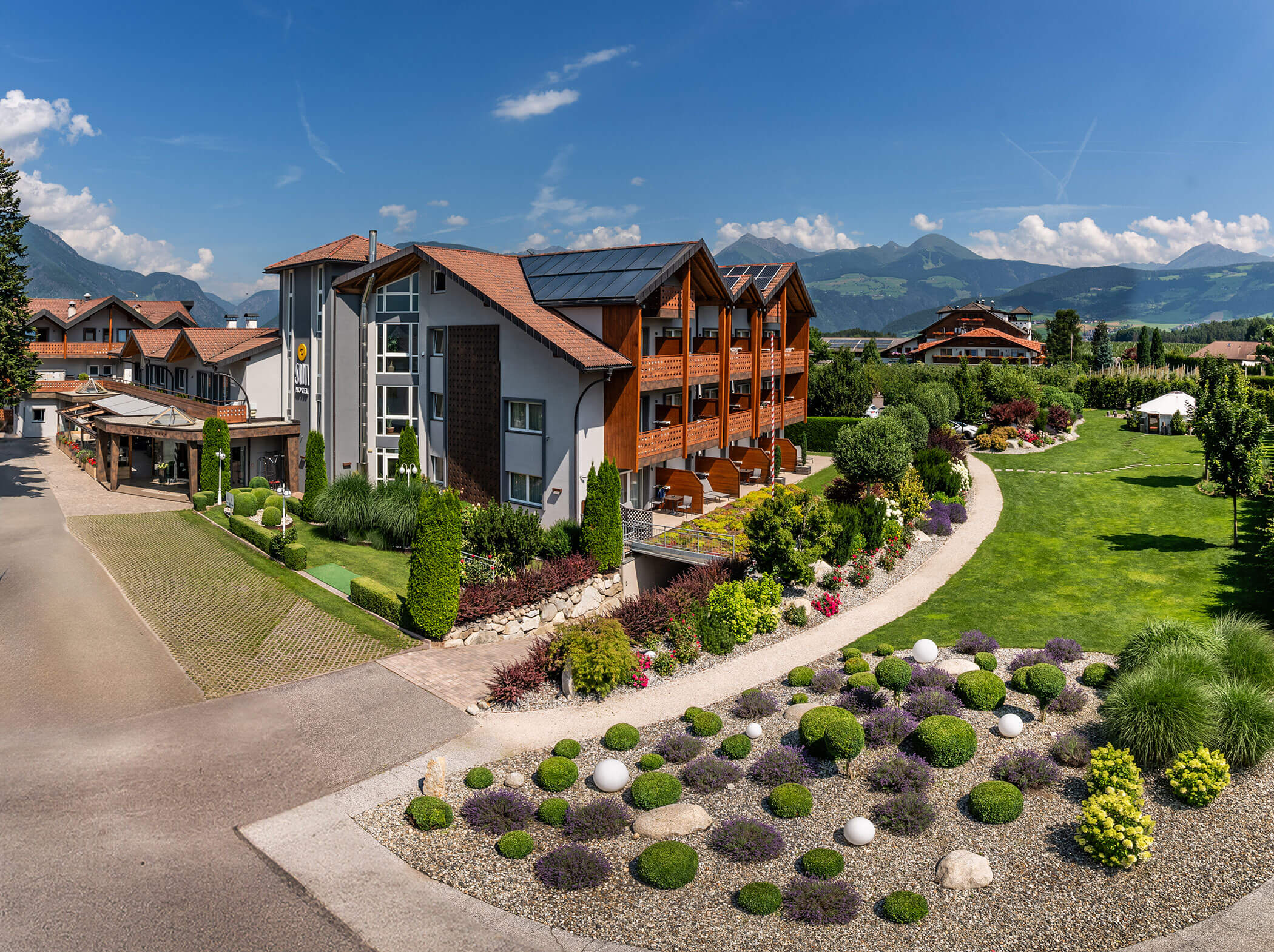 Modern alpine hotel with wood accents, manicured gardens, and mountains in the background under a blue sky. - Hotel Sun