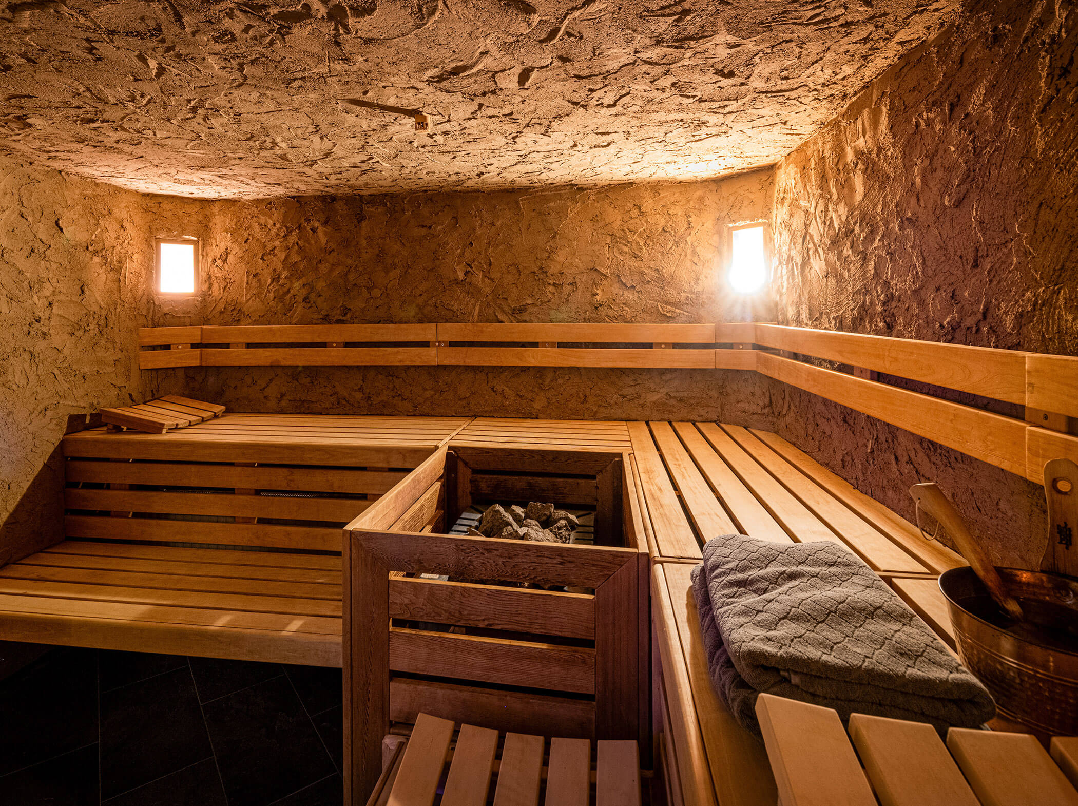 Wood-paneled sauna with benches, heated stones in the center, towels, and soft lighting from two small windows. - Hotel Sun