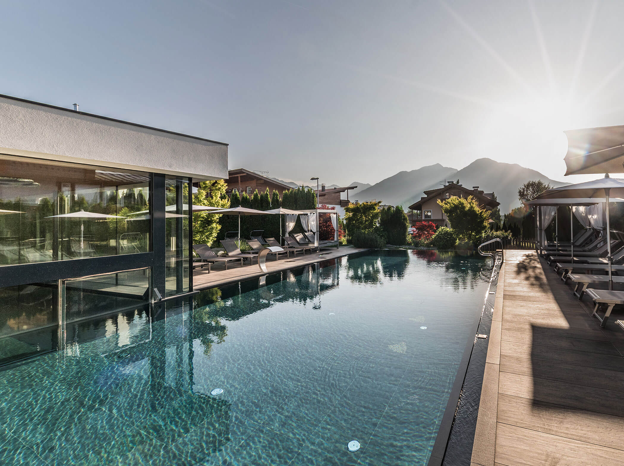 Outdoor pool beside a modern building, with lounge chairs and mountains in the background at sunrise. - Hotel Sun