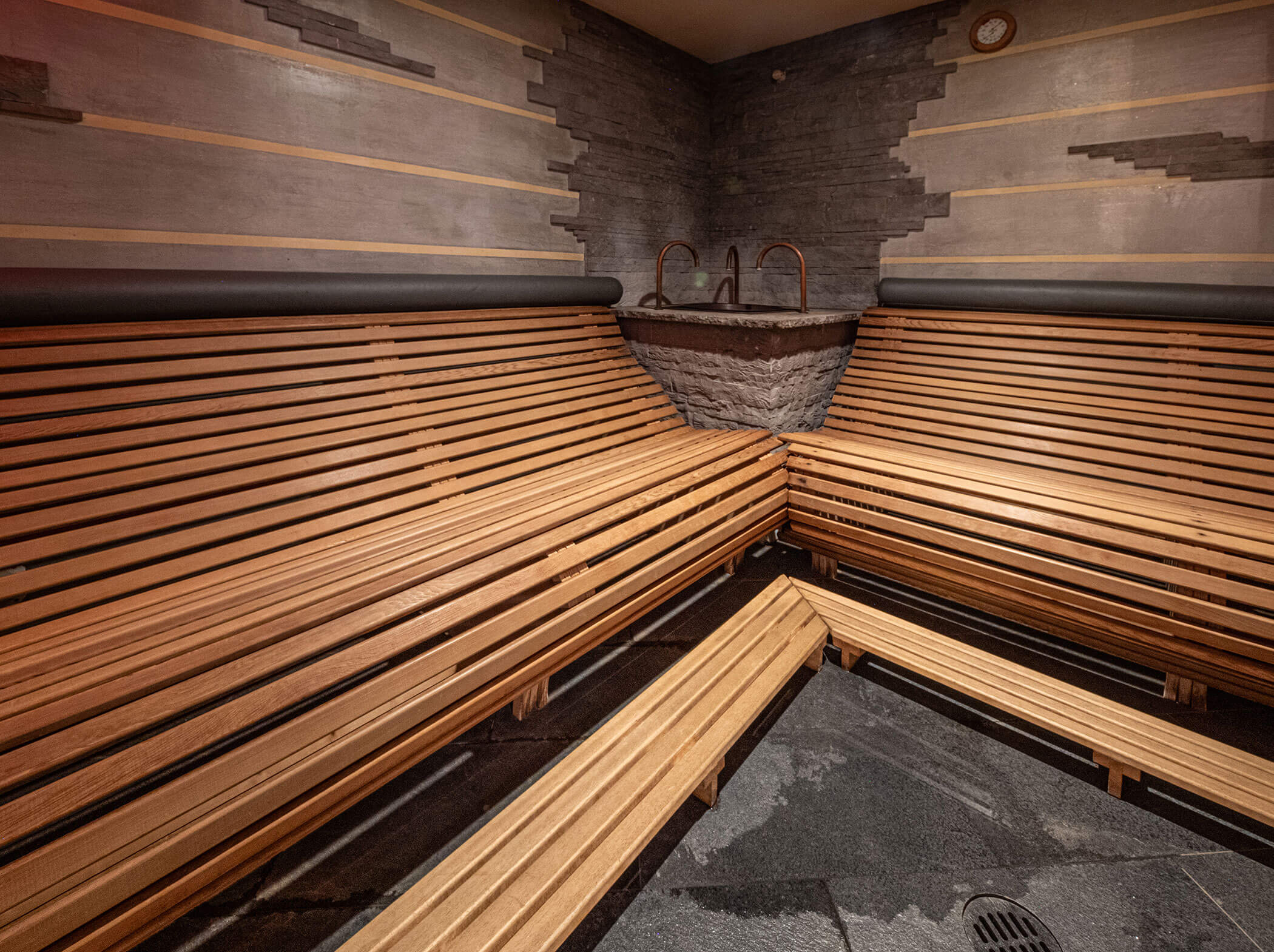 Leere Holzsauna mit gestuften Bänken, Steinwandakzenten und einem Wasserhahn in der Ecke. - Hotel Sun