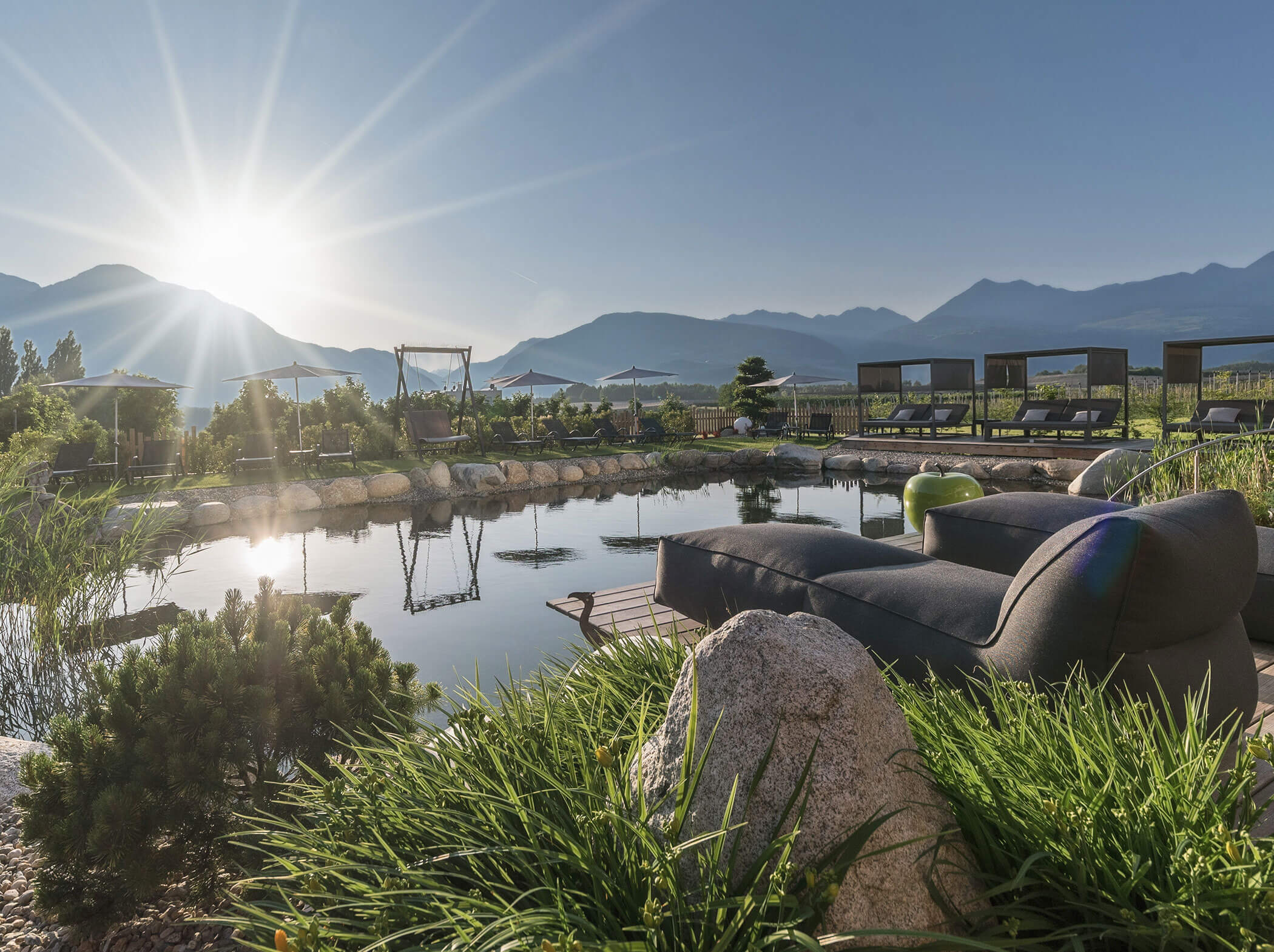 Sunrise over mountains, a tranquil pond, and a modern lounge chair surrounded by lush greenery. - Hotel Sun