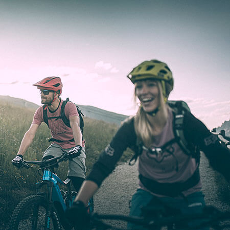 Zwei Personen mit Helmen fahren mit dem Mountainbike auf einem Weg durch grasbewachsene Hügel unter einem bewölkten Himmel. - Hotel Sun