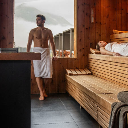 Ein Mann und eine Frau entspannen sich in einer Holzsauna mit einem großen Fenster, das eine Aussicht auf die Berge bietet. - Hotel Sun