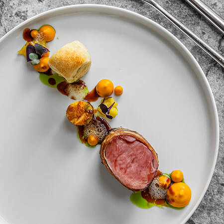 Elegantes Gericht mit aufgeschnittenem Fleisch, Soßentupfen, Brötchen und Beilagen, kunstvoll auf einem weißen Teller angerichtet. - Hotel Sun