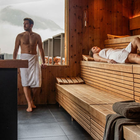 Un uomo e una donna con gli asciugamani si rilassano in una sauna di legno con vista sulle montagne fuori dalla finestra. - Hotel Sun