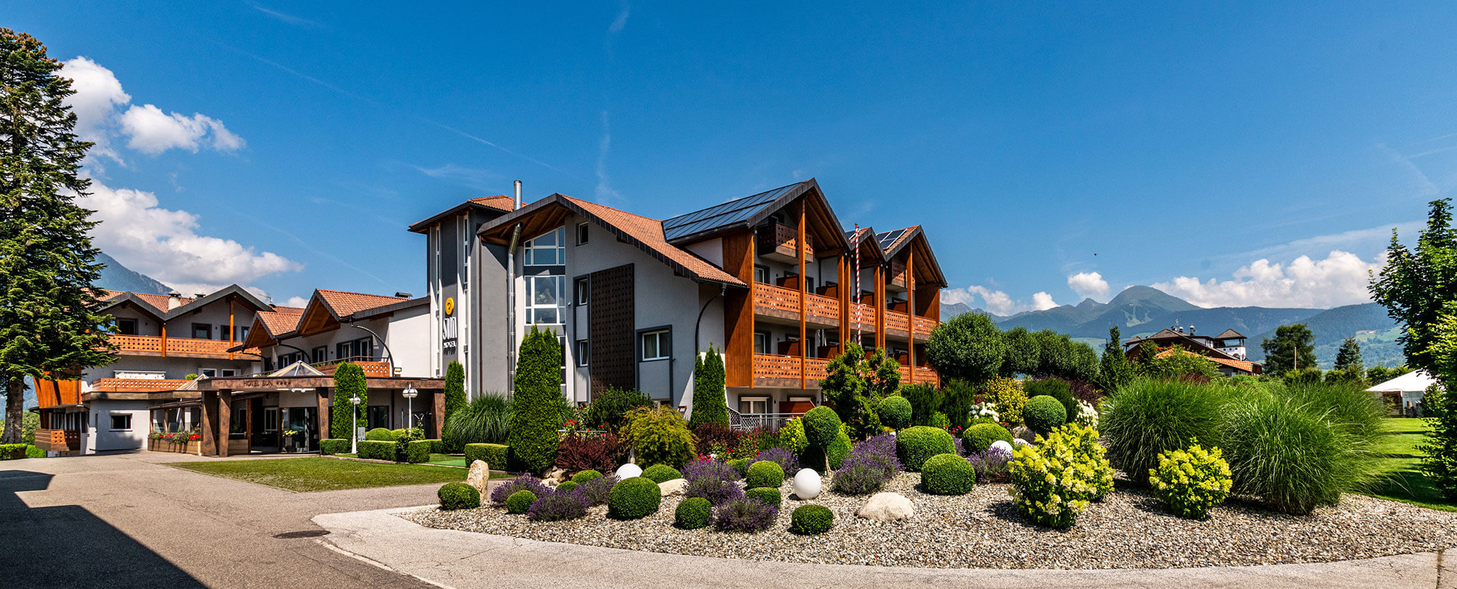 Modern mountain hotel with wooden balconies, lush gardens, and distant mountains under a blue sky. - Hotel Sun