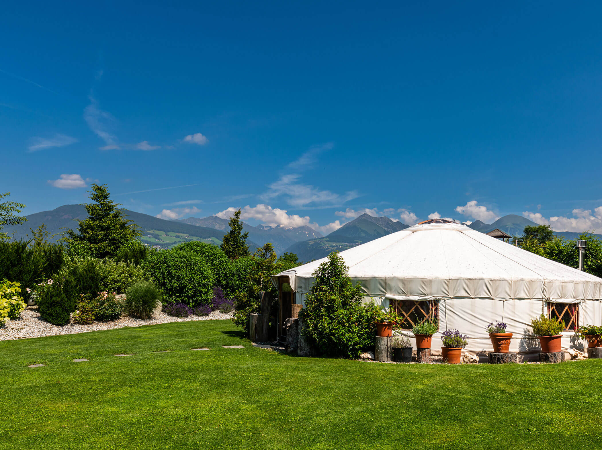 A white yurt sits on green grass with potted plants, trees, and mountains in the background under a blue sky. - Hotel Sun