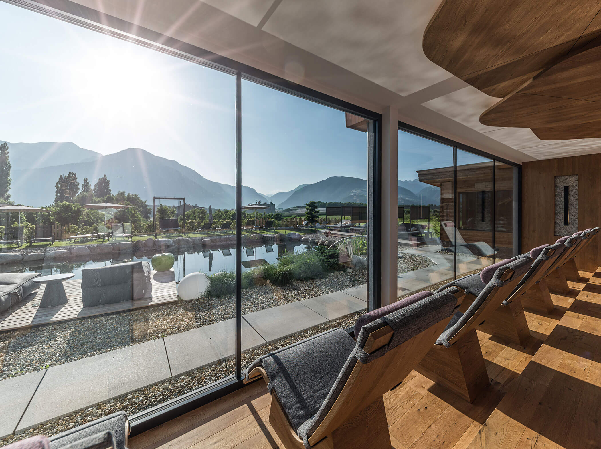 View from inside a modern lounge looking out at a sunny mountain landscape and pond through large glass windows. - Hotel Sun
