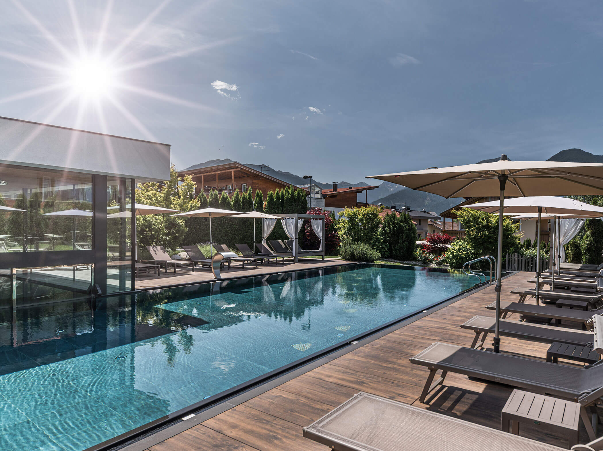 Modern outdoor pool with lounge chairs and umbrellas, sun shining brightly, surrounded by greenery and mountains. - Hotel Sun