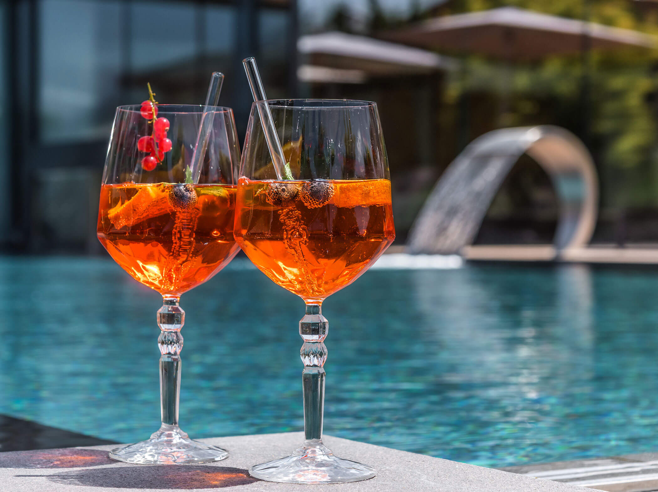 Zwei Orangencocktails mit Eis und Beeren an einem Swimmingpool, mit einem Springbrunnen und Sonnenschirmen im Hintergrund. - Hotel Sun