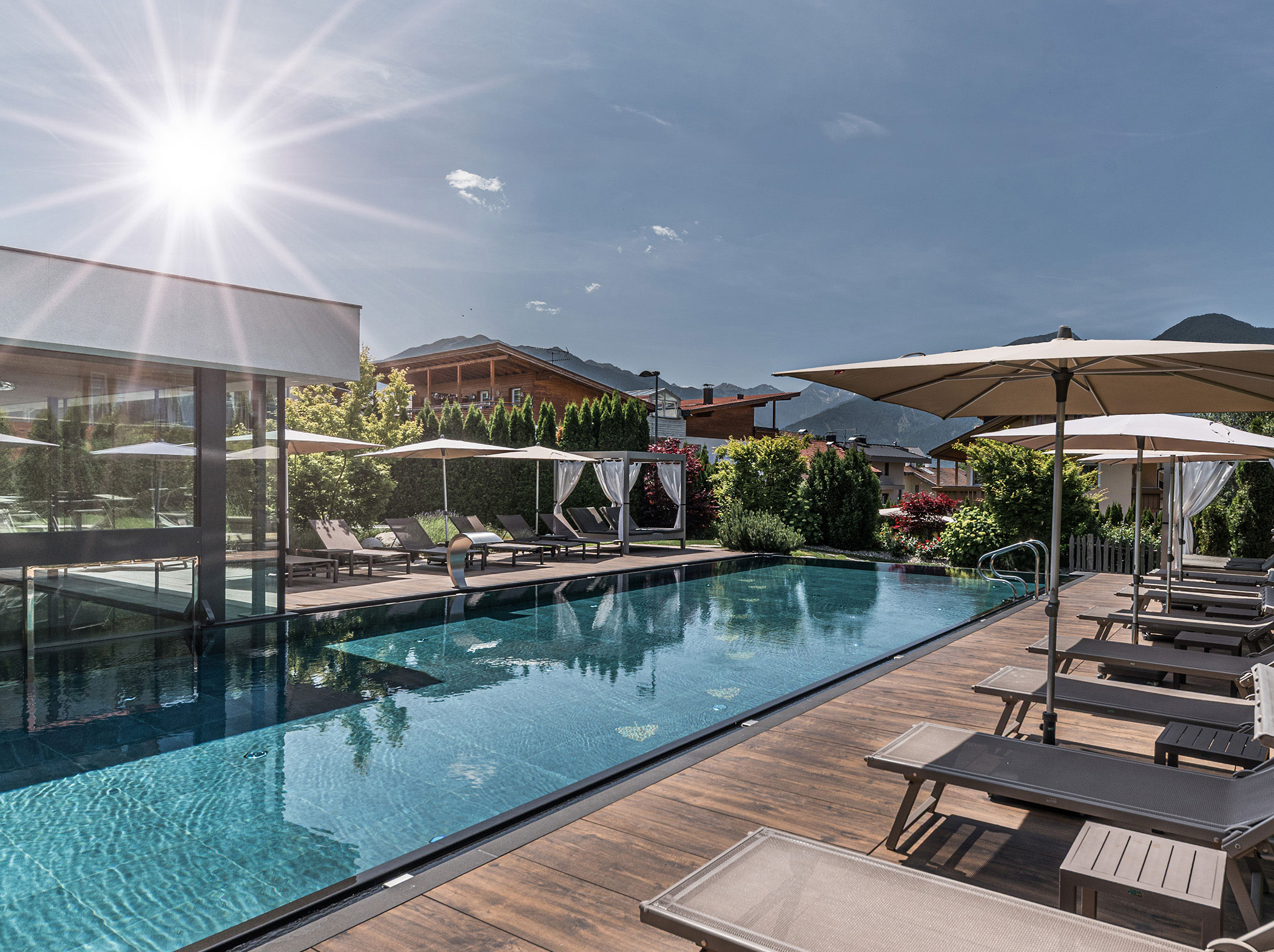 Sonniger Außenpool mit Liegestühlen, Sonnenschirmen und Bergblick neben einem modernen Glasgebäude. - Hotel Sun