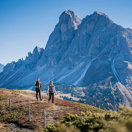 Zwei Personen wandern auf einem Bergpfad mit einem hoch aufragenden, felsigen Gipfel im Hintergrund unter einem klaren Himmel. - Hotel Sun