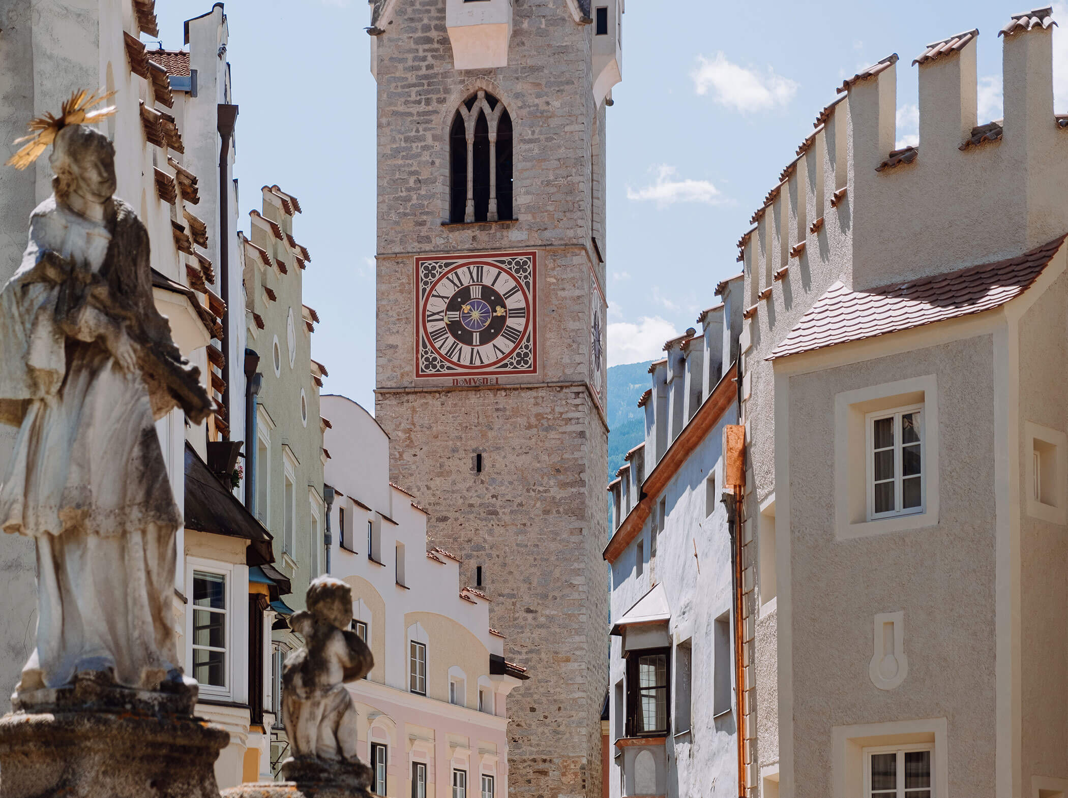 Steinerner Uhrenturm zwischen historischen pastellfarbenen Gebäuden, Statuen im Vordergrund, unter blauem Himmel. - Hotel Sun