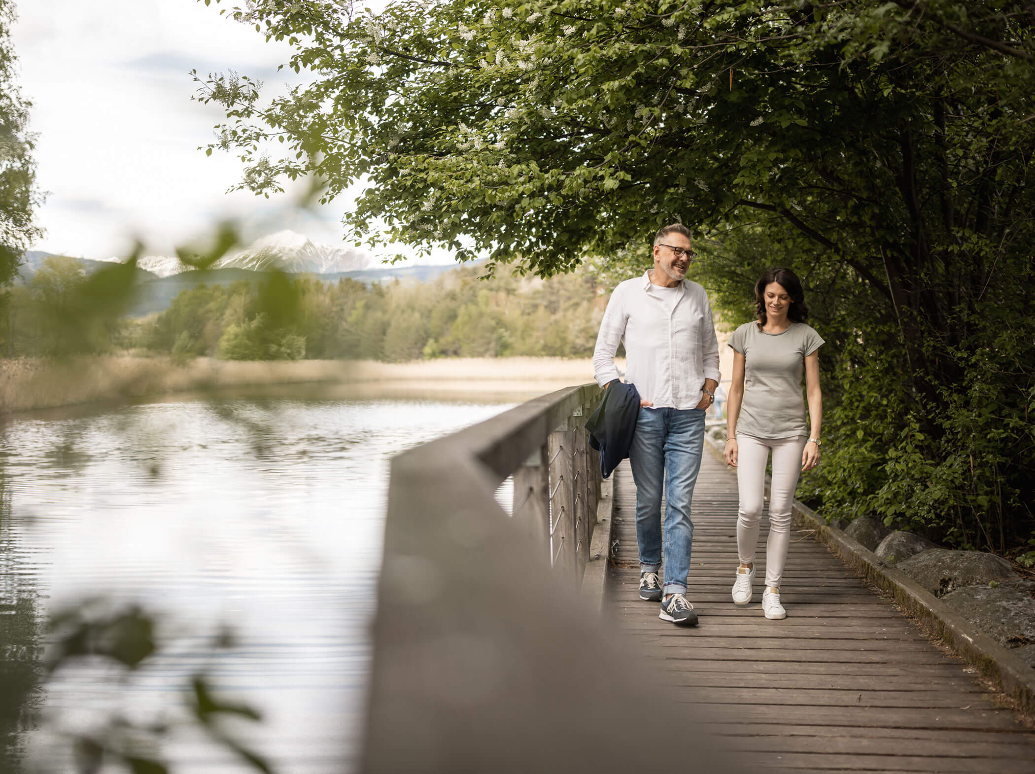 Ein Mann und eine Frau gehen gemeinsam auf einem Holzweg an einem See, umgeben von Bäumen. - Hotel Sun