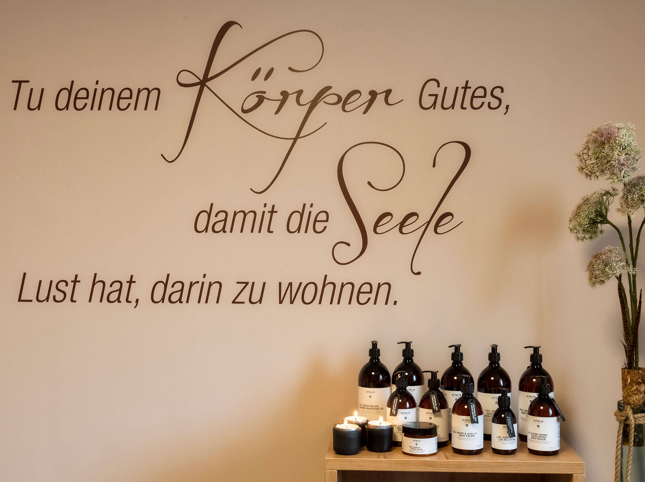 Eine Wand mit deutschem Text, Kerzen, Flaschen auf einem Regal und eine Vase mit Blumen in einer ruhigen, kurähnlichen Umgebung. - Hotel Sun