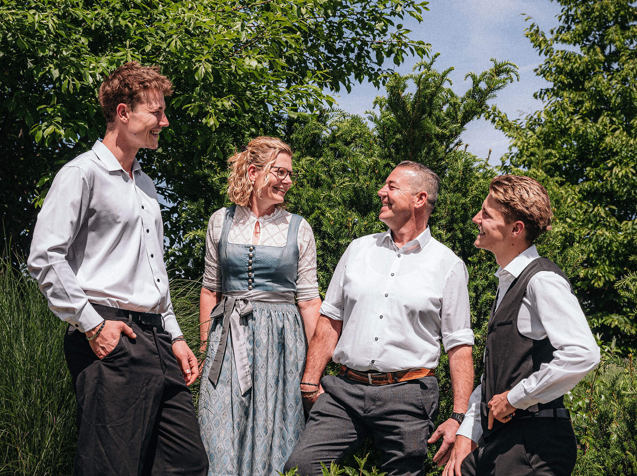 Vier elegant gekleidete Personen stehen im Freien, lächeln und unterhalten sich, während im Hintergrund grüne Bäume zu sehen sind. - Hotel Sun