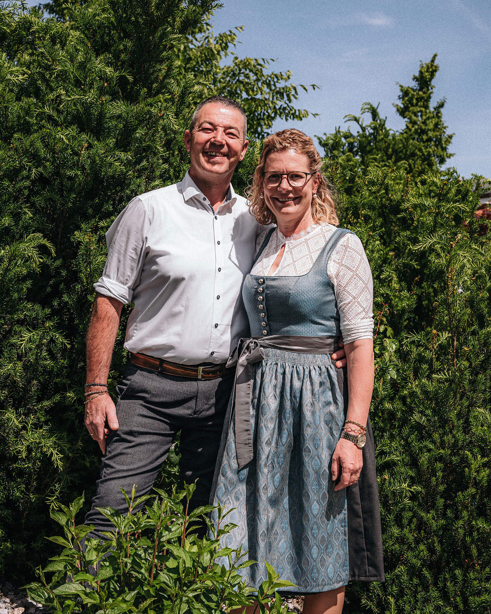 Lächelndes Paar, das im Freien posiert; Mann in weißem Hemd, Frau in traditionellem Kleid, Grünzeug im Hintergrund. - Hotel Sun