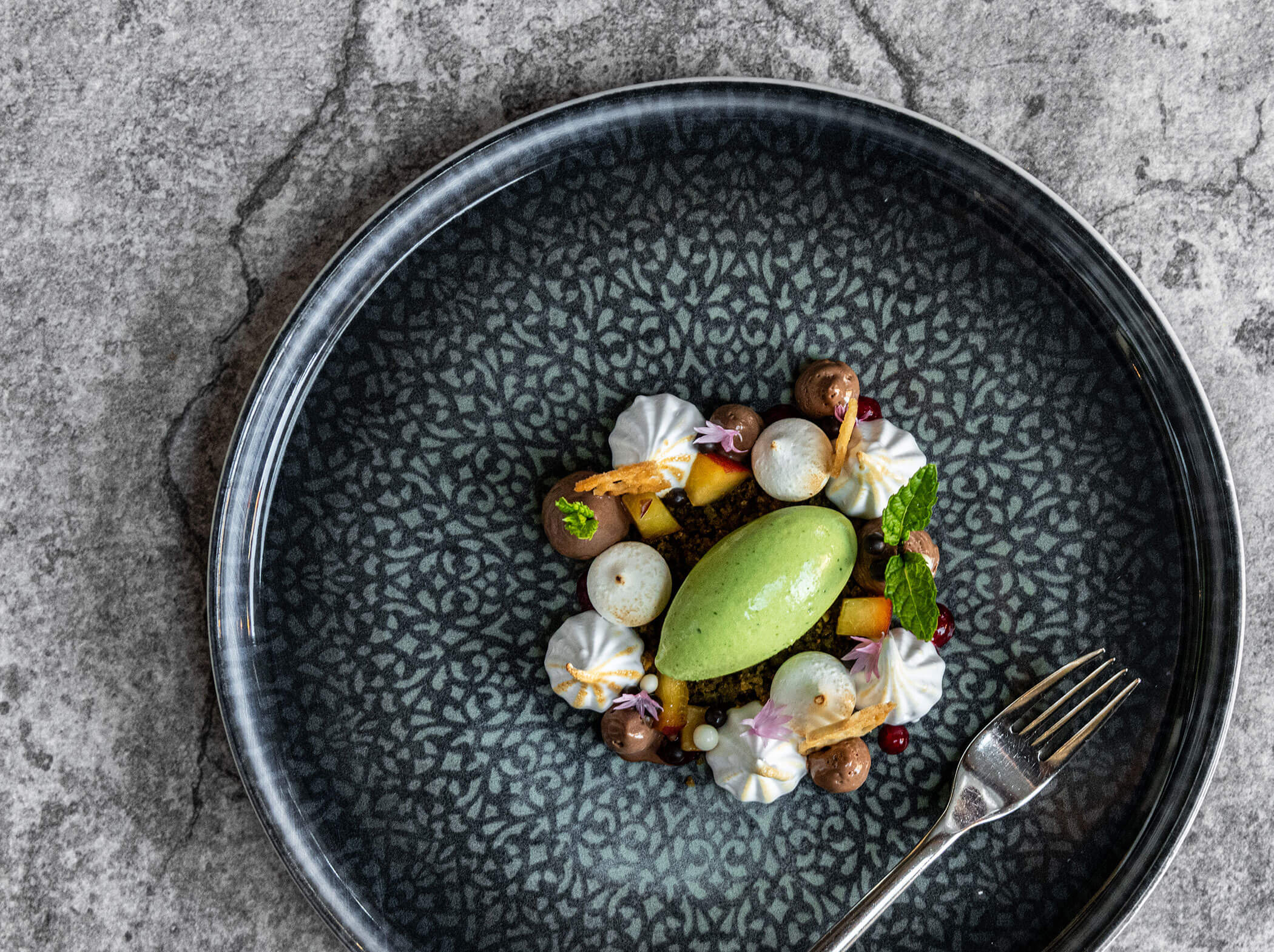 Ein Gourmet-Dessert mit grünem Sorbet, Meringues, Früchten und Minze, kunstvoll angerichtet auf einem gemusterten Teller. - Hotel Sun