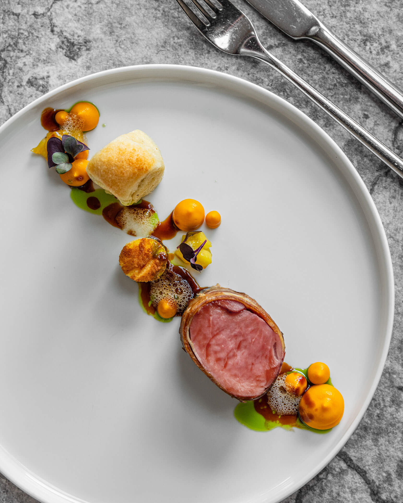 Ein Feinschmeckergericht mit aufgeschnittenem Fleisch, Brot, Saucen, Kräutern und Beilagen auf einem weißen Teller mit Besteck daneben. - Hotel Sun