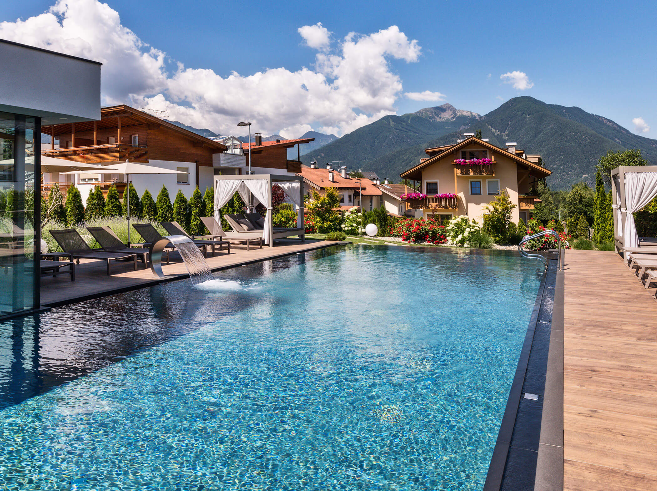 Ein Swimmingpool mit Liegestühlen, Cabanas und Blick auf die Berge unter einem blauen Himmel mit Wolken. - Hotel Sun
