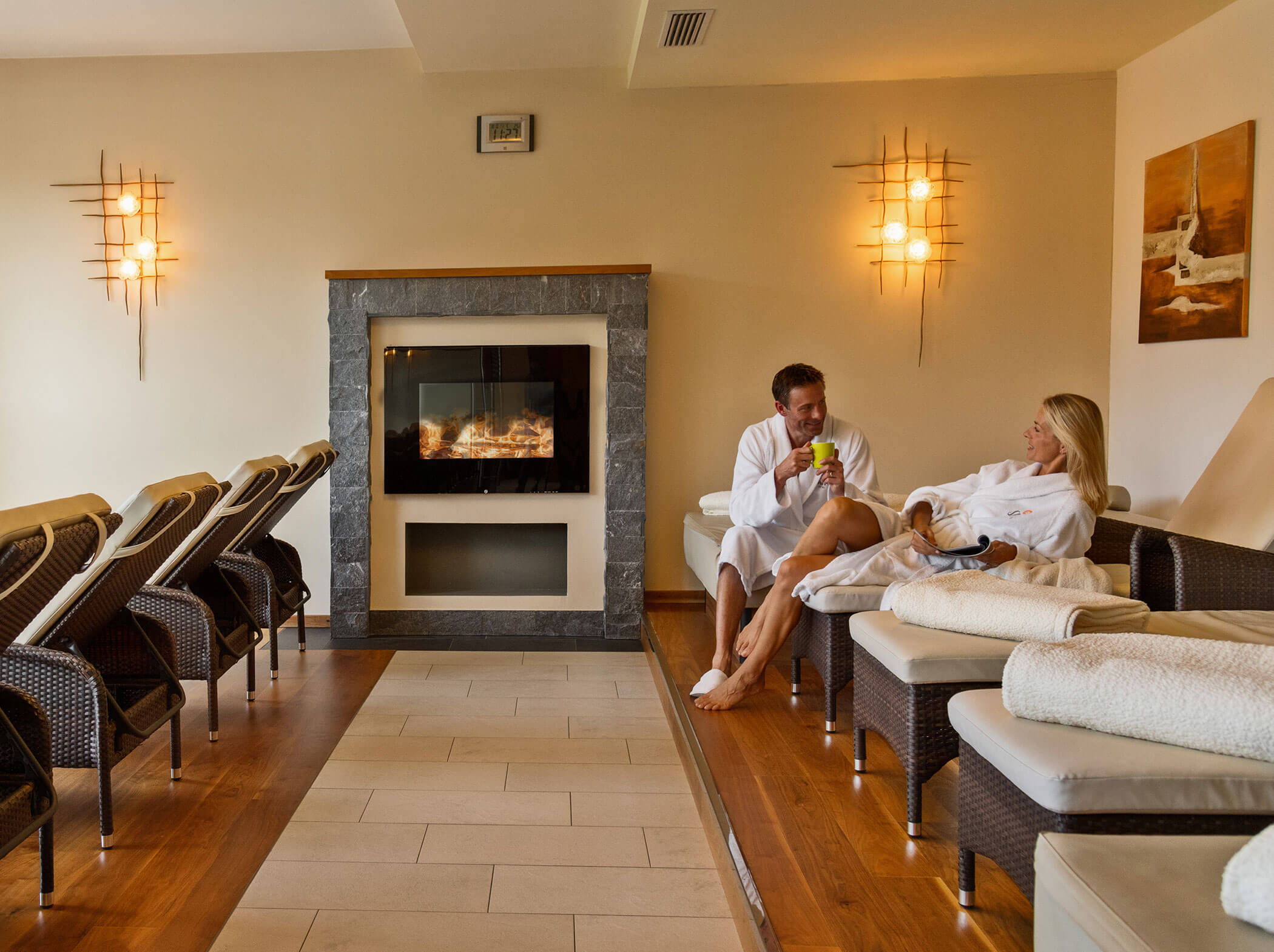 Ein Mann und eine Frau in Bademänteln entspannen sich an einem Kamin in einer gemütlichen Spa-Lounge mit Liegestühlen und warmer Beleuchtung. - Hotel Sun