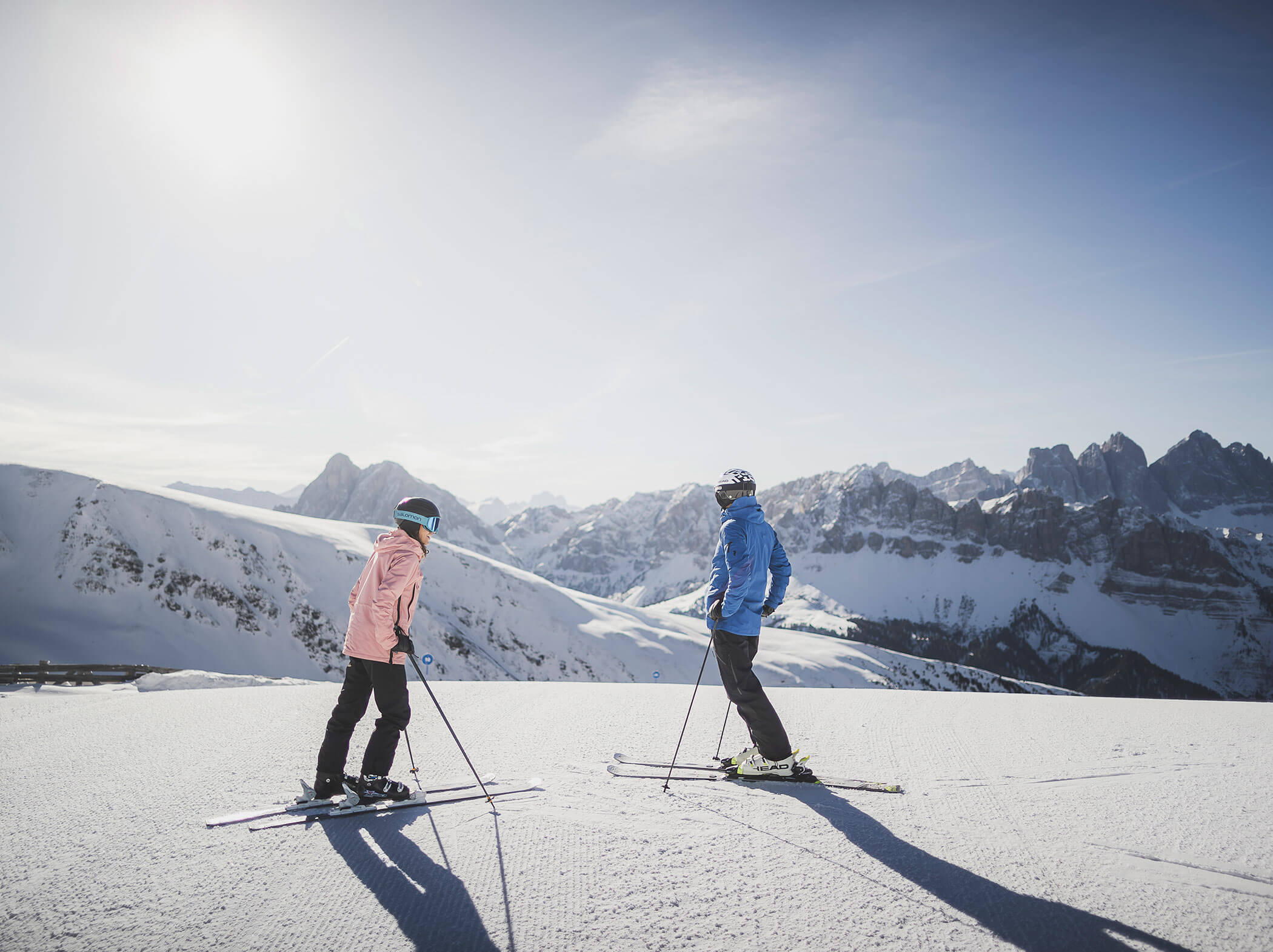 Zwei Personen beim Skifahren auf einem verschneiten Berghang mit einem malerischen Hintergrund von Bergen unter einem klaren Himmel. - Hotel Sun