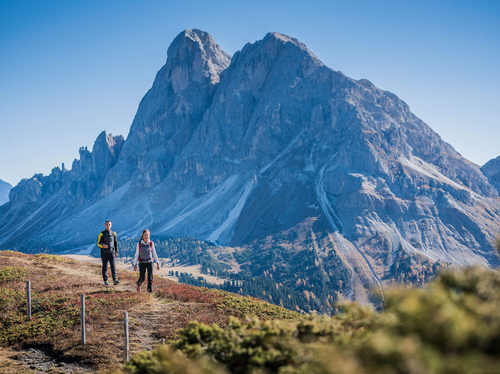 Zwei Personen wandern auf einem Weg mit einem großen felsigen Berg im Hintergrund unter einem klaren blauen Himmel. - Hotel Sun