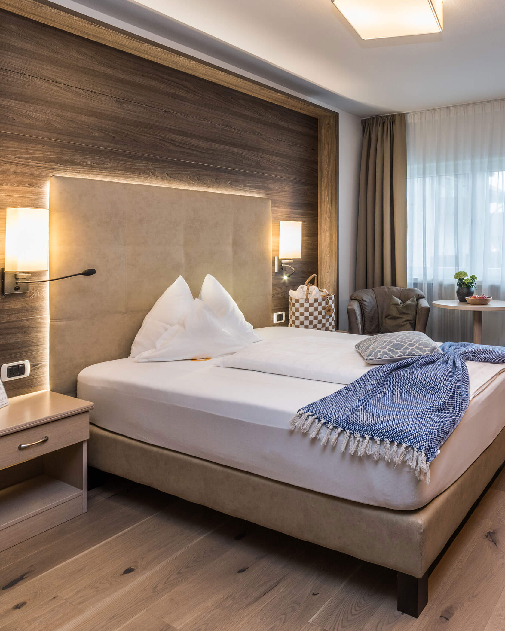 Modernes Hotelzimmer mit einem Doppelbett, sanfter Beleuchtung, einer blauen Decke und einem Fenster mit durchsichtigen Vorhängen. - Hotel Sun