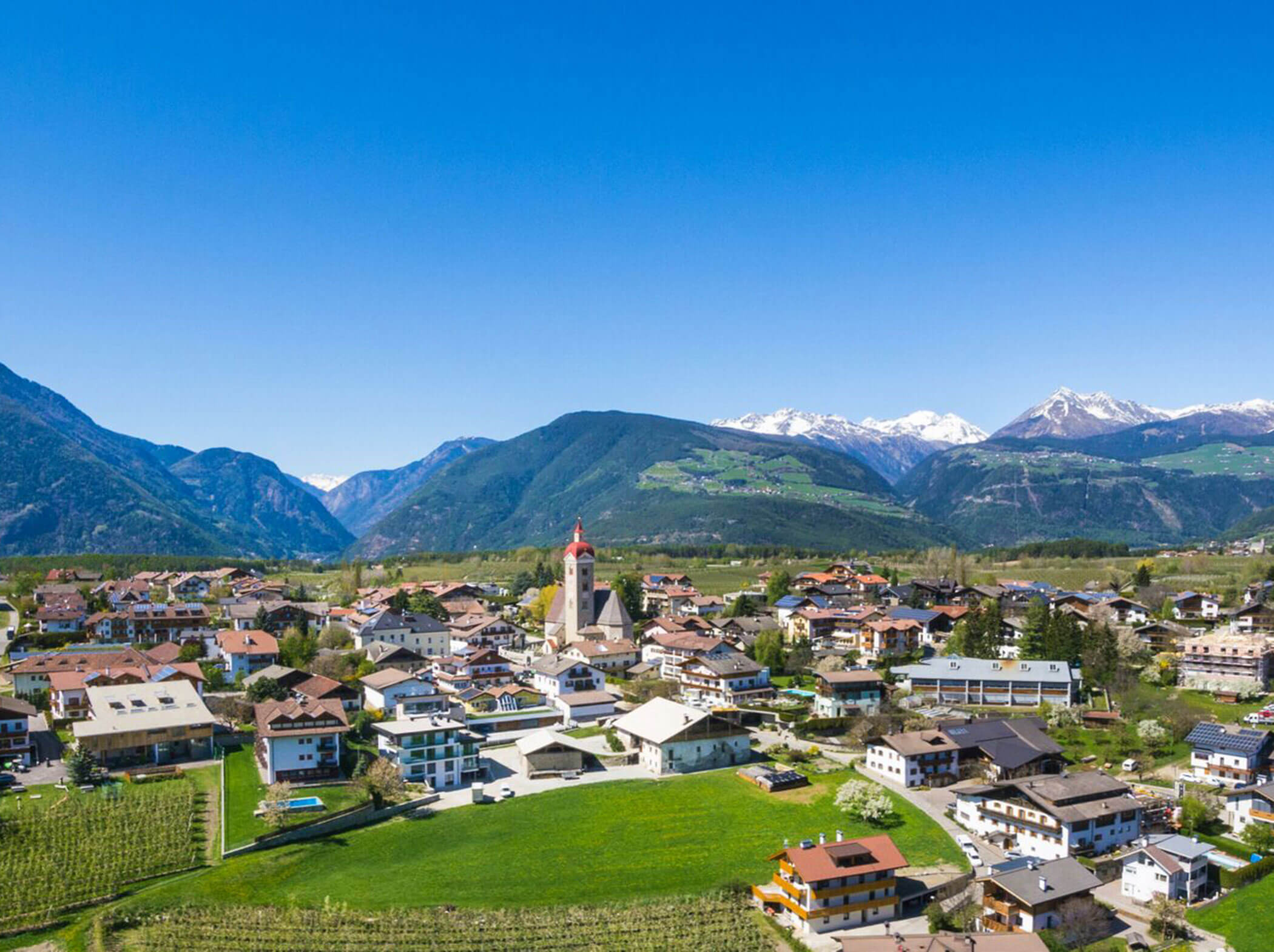 Eine hölzerne Lounge-Bank steht im Gras, umgeben von Bäumen und Bergen im Hintergrund. - Hotel Sun