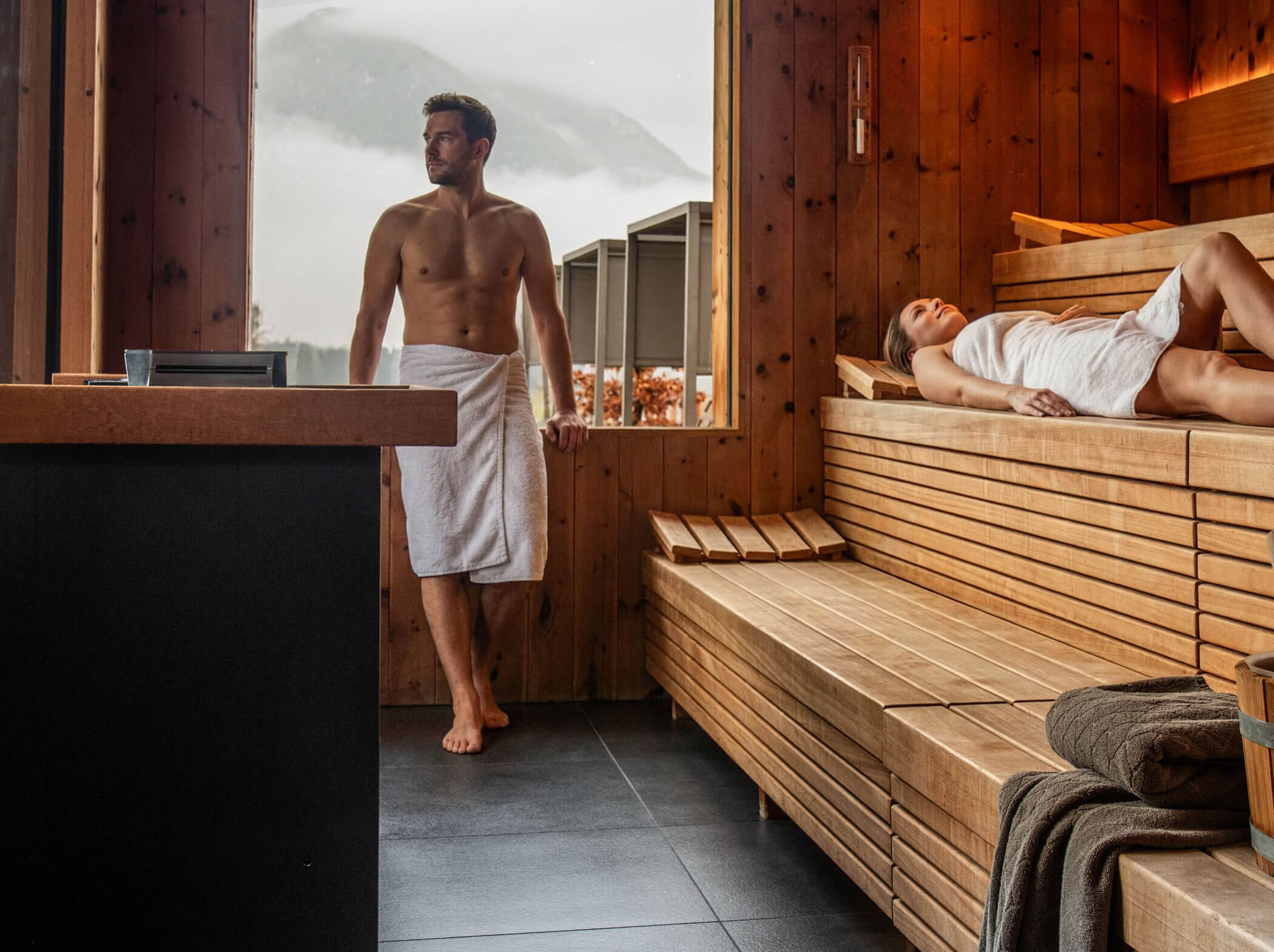 Ein Mann und eine Frau entspannen sich in einer Holzsauna mit Blick auf die Berge durch das Fenster. - Hotel Sun