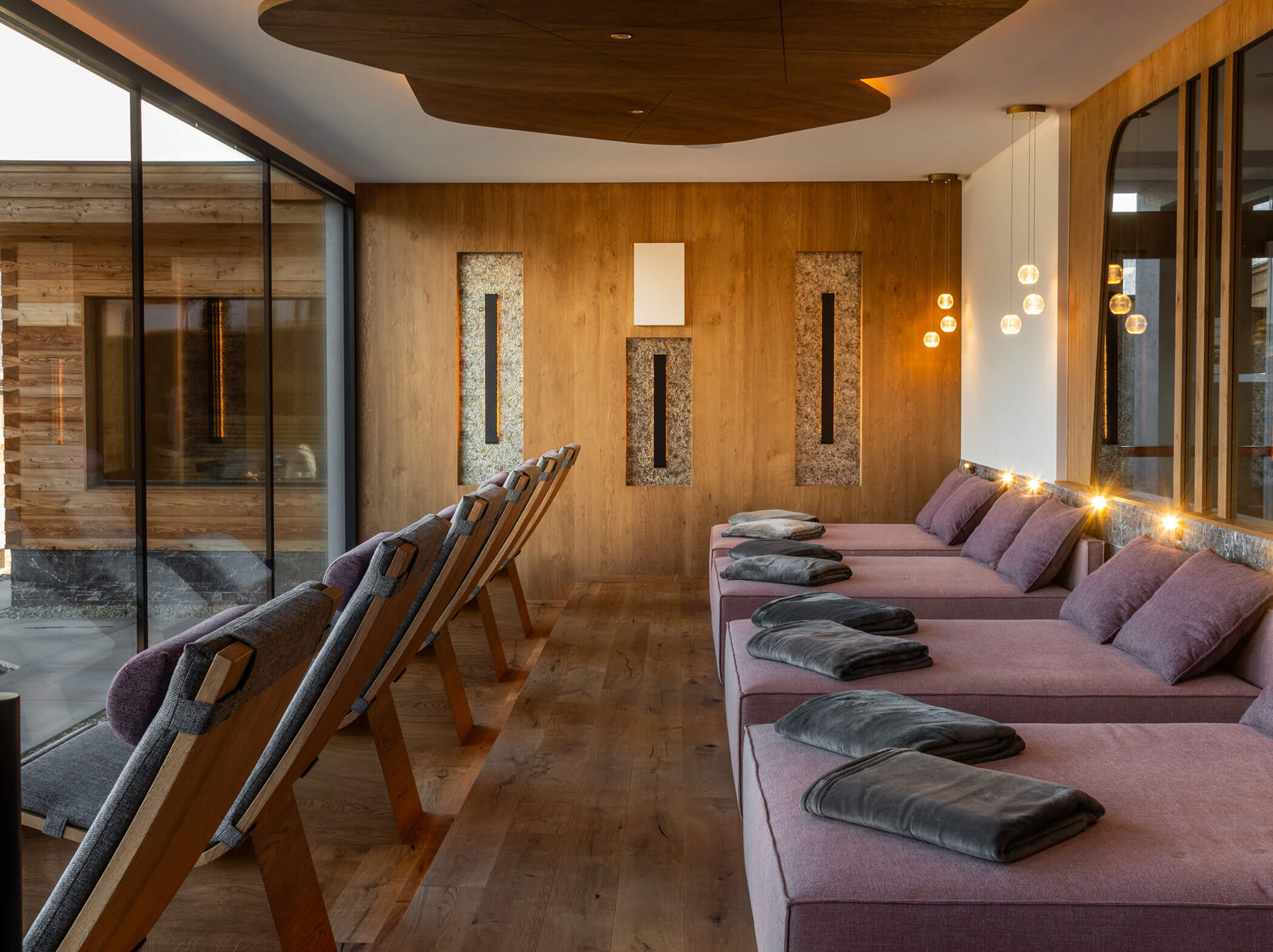 Moderne Spa-Lounge mit lila gepolsterten Sitzen, Holzstühlen, Decken und warmer Beleuchtung an großen Fenstern. - Hotel Sun