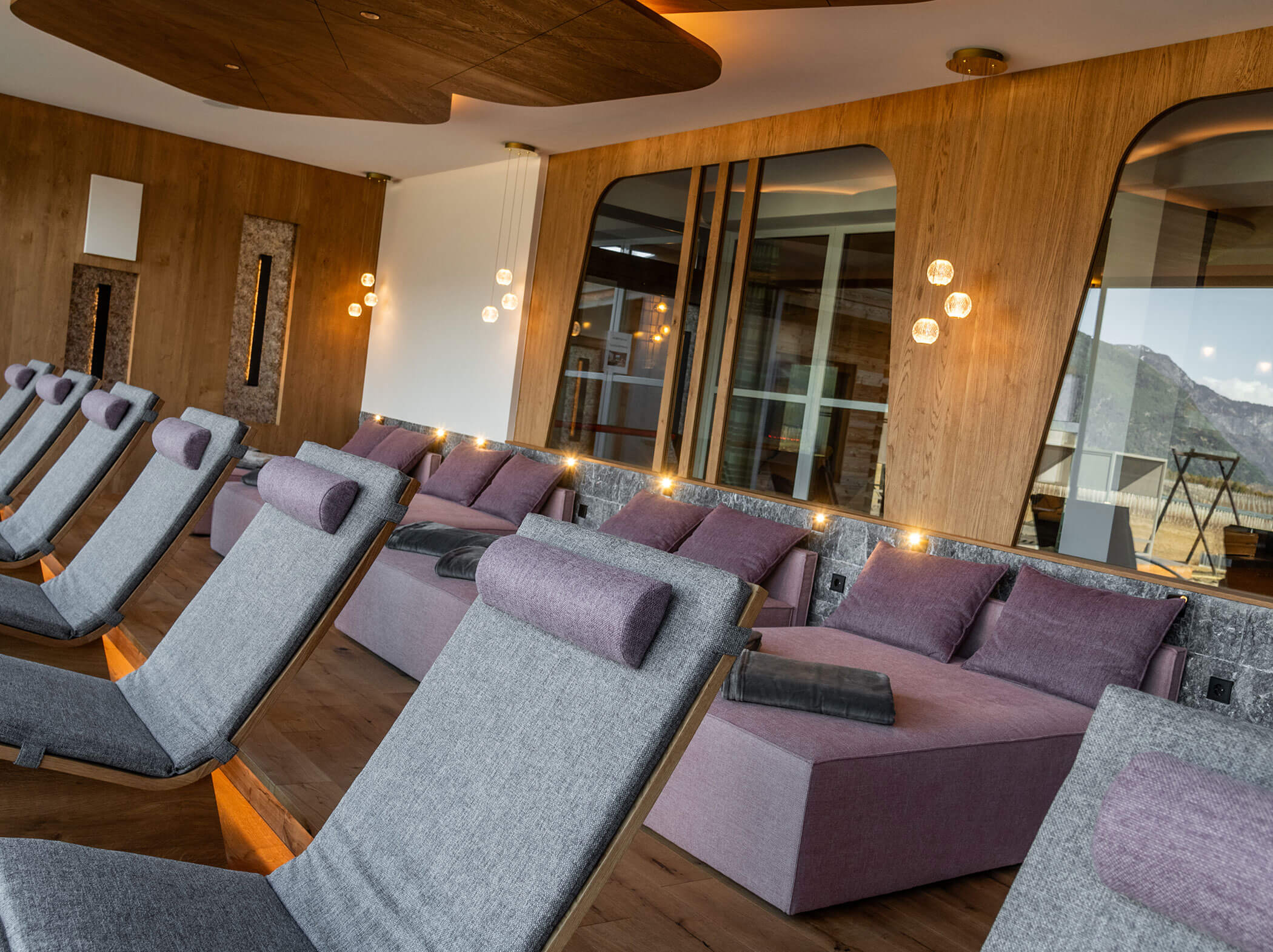 Moderner Lounge-Bereich mit grauen Sesseln, lila Daybeds, Holzwänden und großen Fenstern mit Bergblick. - Hotel Sun