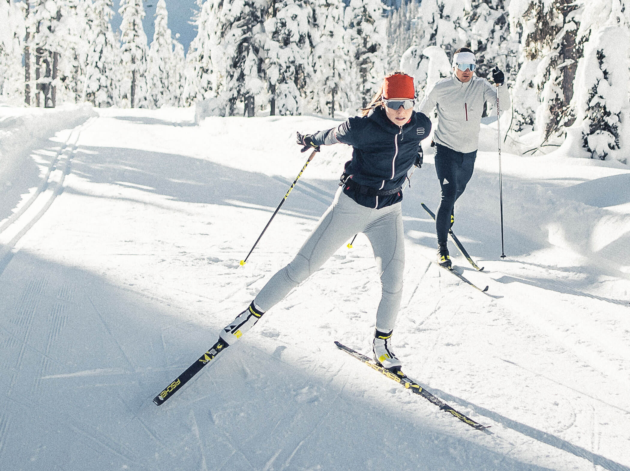 Zwei Personen beim Skilanglauf auf einer verschneiten Loipe, umgeben von schneebedeckten Bäumen und hellem Sonnenlicht. - Hotel Sun