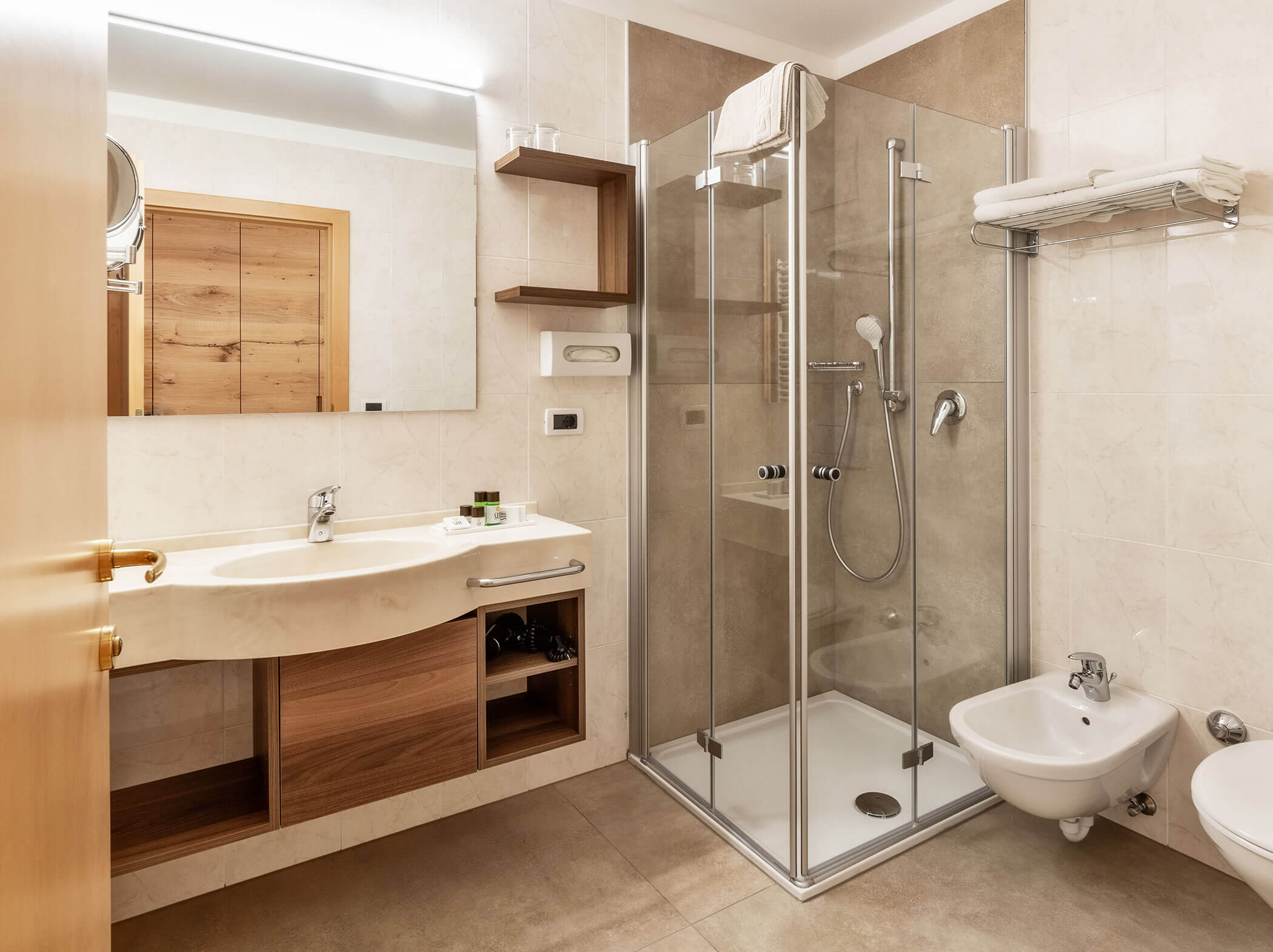 Modernes Badezimmer mit Glasdusche, Bidet, Waschbecken, Holzschränken und Handtüchern, die ordentlich in Regalen angeordnet sind. - Hotel Sun